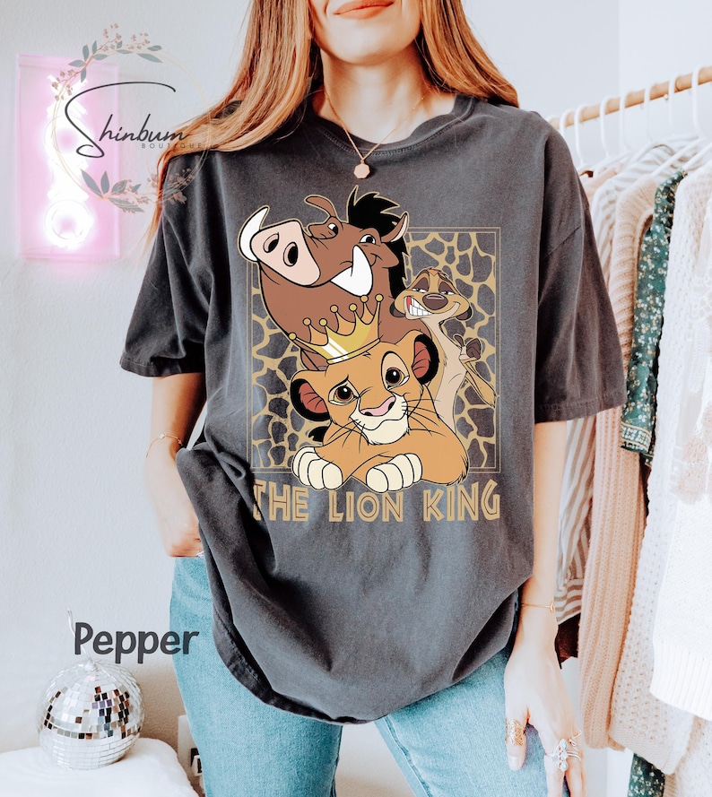 May include: Dark gray t-shirt featuring a graphic of characters from The Lion King, including Simba wearing a crown, Pumbaa, and Timon. The text "THE LION KING" is printed below the characters. The shirt has a relaxed fit.