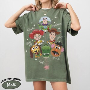 Puede incluir: Camiseta Comfort Colors verde musgo con un gr&aacute;fico de los personajes de Toy Story: Buzz Lightyear, Woody, Jessie, Slinky Dog, Rex y Mr. Potato Head. La camiseta tiene el texto "Jessie", "Woody" y "Potato".