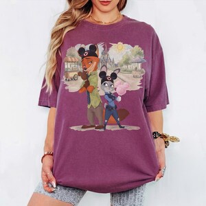 Puede incluir: Una camiseta morada con una ilustraci&oacute;n de dibujos animados de Nick Wilde y Judy Hopps de Zootopia, con orejas de Mickey Mouse. El fondo representa una escena de parque con un castillo y un puesto de comida.
