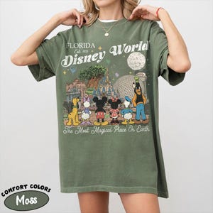 May include: Moss green t-shirt with a Disney World graphic. The design includes Mickey Mouse, Minnie Mouse, and Goofy, with the text "Florida Disney World Est. 1971" and "The Most Magical Place On Earth". The shirt is a Comfort Colors brand.