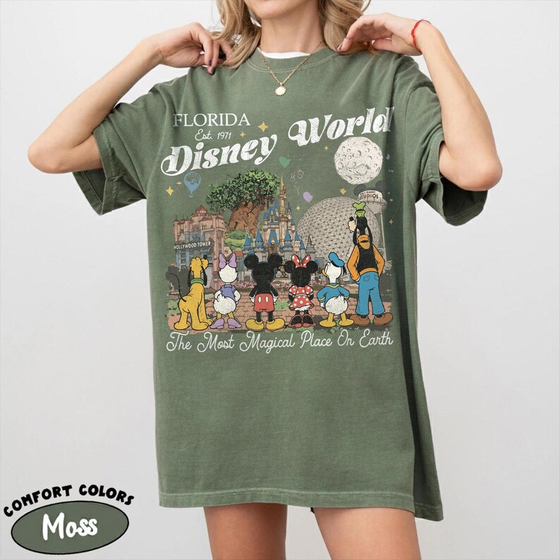 May include: Moss green t-shirt with a Disney World graphic. The design includes Mickey Mouse, Minnie Mouse, and Goofy, with the text "Florida Disney World Est. 1971" and "The Most Magical Place On Earth". The shirt is a Comfort Colors brand.