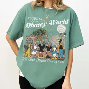 May include: Green t-shirt featuring a Disney World graphic. The design includes the text "Florida Est. 1971 Disney World" and cartoon characters such as Mickey and Minnie Mouse, Donald Duck, and Goofy.