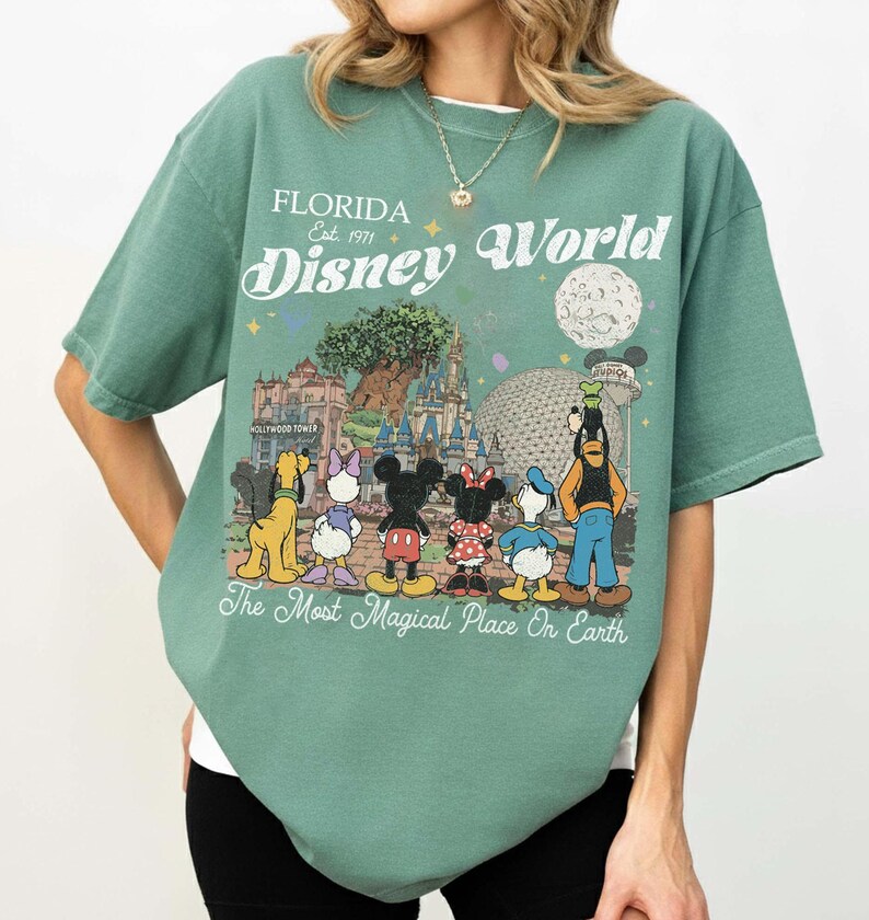 May include: Green t-shirt featuring a Disney World graphic. The design includes the text "Florida Est. 1971 Disney World" and cartoon characters such as Mickey and Minnie Mouse, Donald Duck, and Goofy.
