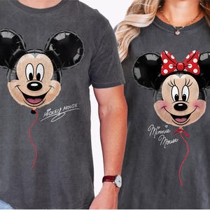 K&ouml;nnte beinhalten: Zwei anthrazitfarbene T-Shirts mit Mickey- und Minnie-Mouse-Ballon-Designs. Mickeys Ballon hat ein l&auml;chelndes Gesicht und den Text "Mickey Mouse". Minnies Ballon hat eine rote Schleife und den Text "Minnie Mouse".