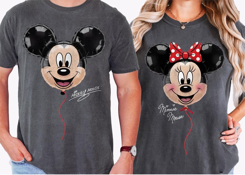 K&ouml;nnte beinhalten: Zwei anthrazitfarbene T-Shirts mit Mickey- und Minnie-Mouse-Ballon-Designs. Mickeys Ballon hat ein l&auml;chelndes Gesicht und den Text "Mickey Mouse". Minnies Ballon hat eine rote Schleife und den Text "Minnie Mouse".
