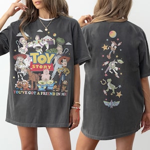 Op de afbeelding: Een antracietgrijs T-shirt met korte mouwen en een Toy Story-opdruk op de voorkant met personages zoals Woody, Buzz Lightyear en Jessie, plus de tekst "TOY STORY" en "YOU'VE GOT A FRIEND IN ME". De achterkant toont een verticaal ontwerp.