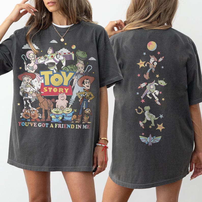 Op de afbeelding: Een antracietgrijs T-shirt met korte mouwen en een Toy Story-opdruk op de voorkant met personages zoals Woody, Buzz Lightyear en Jessie, plus de tekst "TOY STORY" en "YOU'VE GOT A FRIEND IN ME". De achterkant toont een verticaal ontwerp.