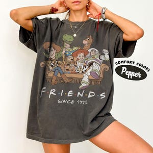 May include: A charcoal gray t-shirt featuring a graphic print of various animated characters sitting on a couch. Below the characters, the text reads "FRIENDS SINCE 1995". A circular graphic on the right side displays the text "COMFORT COLORS Pepper".