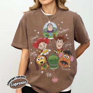 Puede incluir: Camiseta color espresso con un gr&aacute;fico de personajes de Toy Story, incluyendo a Woody, Buzz Lightyear y Jessie. El dise&ntilde;o incluye los textos "Buzz Lightyear", "Jessie", "Woody", "Slinky Dog", "Rex" y "Potato". La camiseta tambi&eacute;n tiene el texto "COMFORT COLORS" y "Espresso".