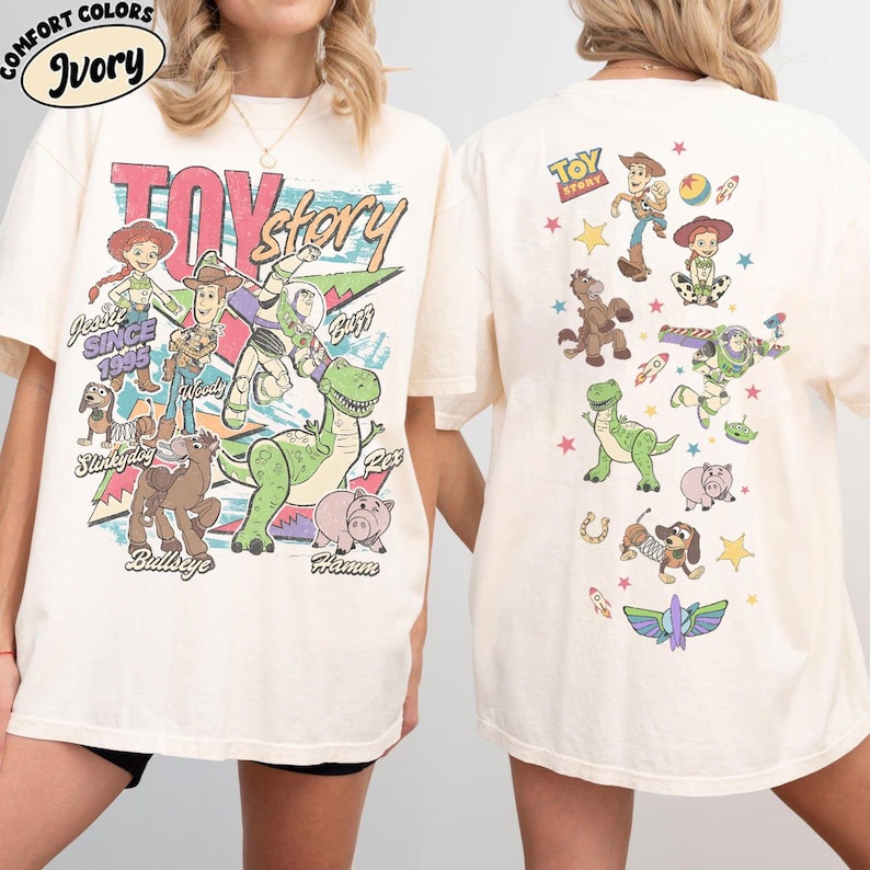 May include: Ivory-colored t-shirt featuring a colorful graphic of Toy Story characters. The front design includes the words "Toy Story" and various characters. The back of the shirt has additional character illustrations. The shirt has short sleeves.