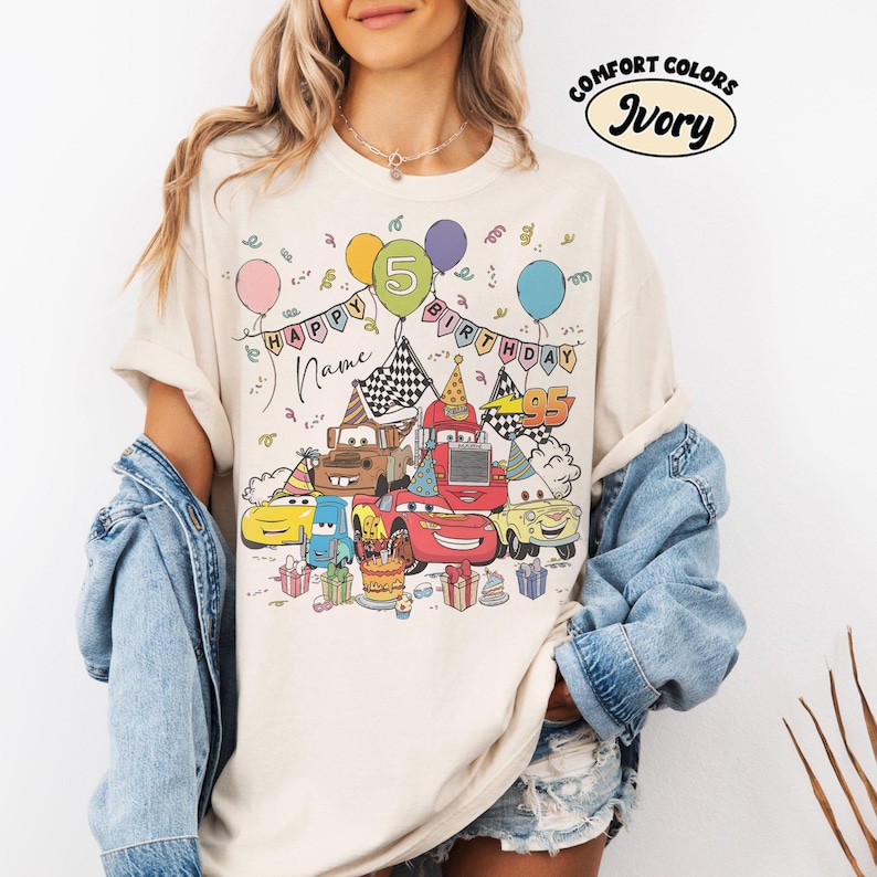 K&ouml;nnte beinhalten: Elfenbeinfarbenes T-Shirt mit einem farbenfrohen Cartoon-Design von Cars-Charakteren, die einen Geburtstag feiern. Das Design umfasst Luftballons, eine Geburtstagstorte und den Text "HAPPY BIRTHDAY" und "5". Das Shirt ist aus Comfort Colors Material.