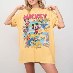 Peut inclure: T-shirt jaune avec un imprim&eacute; de Mickey Mouse et ses amis. Le motif comprend Mickey, Minnie, Donald Duck, Daisy Duck, Dingo et Pluto, avec le texte "Mickey & Friends Since 1928".