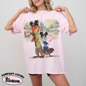 Puede incluir: Camiseta rosa claro con una ilustraci&oacute;n de dibujos animados de Nick Wilde y Judy Hopps de Zootopia, con orejas de Mickey Mouse. El dise&ntilde;o incluye una escena de parque con un castillo y algod&oacute;n de az&uacute;car. La camiseta tiene la inscripci&oacute;n "Comfort Colors Blossom".