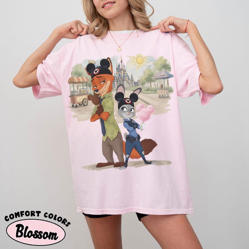 Puede incluir: Camiseta rosa claro con una ilustraci&oacute;n de dibujos animados de Nick Wilde y Judy Hopps de Zootopia, con orejas de Mickey Mouse. El dise&ntilde;o incluye una escena de parque con un castillo y algod&oacute;n de az&uacute;car. La camiseta tiene la inscripci&oacute;n "Comfort Colors Blossom".