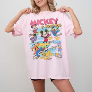 Peut inclure: T-shirt rose clair avec l'inscription "Mickey & Friends" et divers personnages de dessins anim&eacute;s, dont Mickey Mouse, Minnie Mouse, Donald Duck et Dingo. Le t-shirt porte &eacute;galement l'inscription "Since 1928".