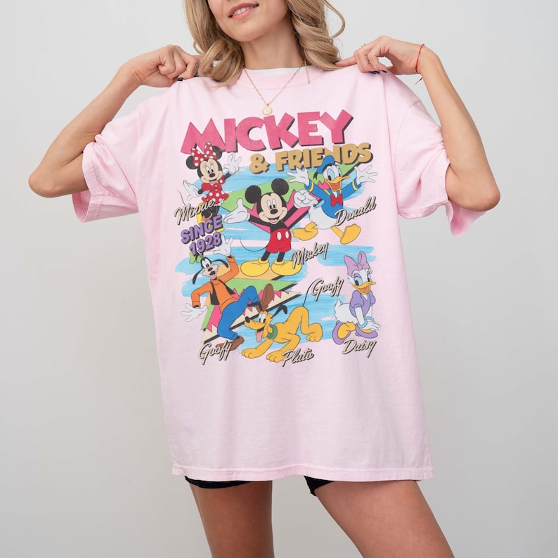 Peut inclure: T-shirt rose clair avec l'inscription "Mickey & Friends" et divers personnages de dessins anim&eacute;s, dont Mickey Mouse, Minnie Mouse, Donald Duck et Dingo. Le t-shirt porte &eacute;galement l'inscription "Since 1928".