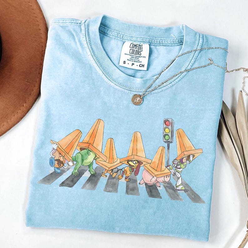 May include: Light blue Comfort Colors t-shirt featuring a whimsical illustration of cartoon characters crossing a crosswalk, each wearing an orange traffic cone. A gold necklace with a pendant rests on the shirt.