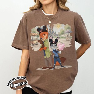 Puede incluir: Camiseta marr&oacute;n con una ilustraci&oacute;n de dibujos animados de dos personajes frente a un castillo y carritos de comida. Los personajes son un zorro y un conejo, ambos con orejas de Mickey Mouse. La camiseta tiene la inscripci&oacute;n "COMFORT COLORS Espresso".