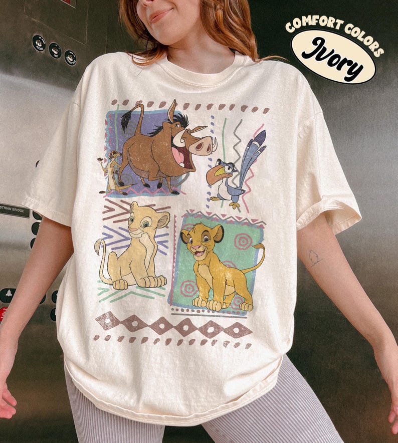 May include: Ivory-colored t-shirt featuring a colorful graphic of characters from The Lion King. The design includes Simba, Timon, Pumbaa, and Zazu. The shirt has a relaxed fit and the words "Comfort Colors" and "Ivory" are printed on the top right.