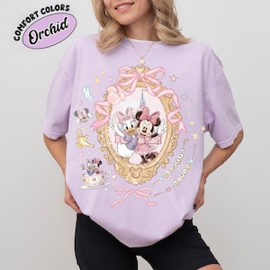 May include: A light purple t-shirt featuring a framed illustration of Minnie Mouse and Daisy Duck. The shirt has a gold ornate frame with pink bows and hearts. The text "COMFORT COLORS Orchid" is in a circular design at the top left.
