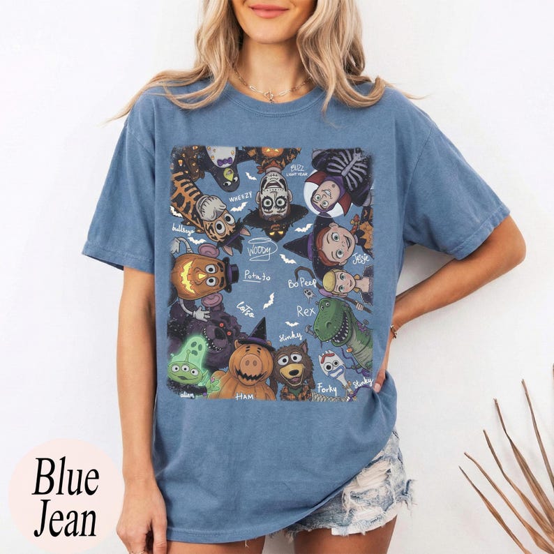 May include: A blue short-sleeve t-shirt featuring a rectangular graphic print of various animated characters dressed in Halloween costumes. The characters include a pumpkin-headed pig, a green alien, a dinosaur, and others, with small white bats and handwritten names surrounding them.