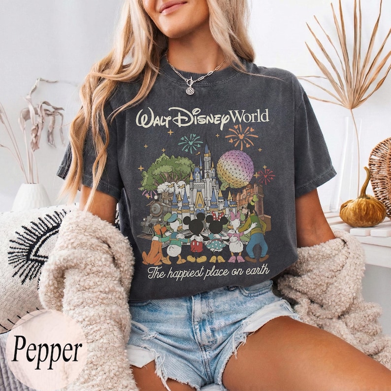 Puede incluir: Camiseta gris oscuro con un gr&aacute;fico colorido de Walt Disney World con el texto "Walt Disney World" y "The happiest place on earth". El gr&aacute;fico incluye personajes de dibujos animados, un castillo y fuegos artificiales.