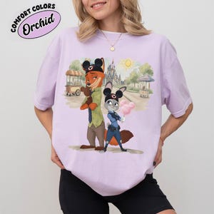 Puede incluir: Camiseta lila claro con una ilustraci&oacute;n de dibujos animados de Nick Wilde y Judy Hopps de Zootopia. La imagen incluye una escena de parque con un castillo y algod&oacute;n de az&uacute;car. El texto "COMFORT COLORS Orchid" est&aacute; impreso en la parte superior izquierda.