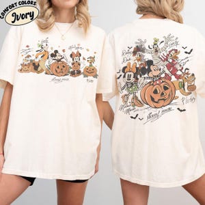 May include: Ivory-colored t-shirt featuring a Halloween-themed graphic of Disney characters, including Mickey Mouse, Minnie Mouse, and Goofy, with pumpkins. The front and back of the shirt are shown. The text "Comfort Colors" and "Ivory" are at the top left.