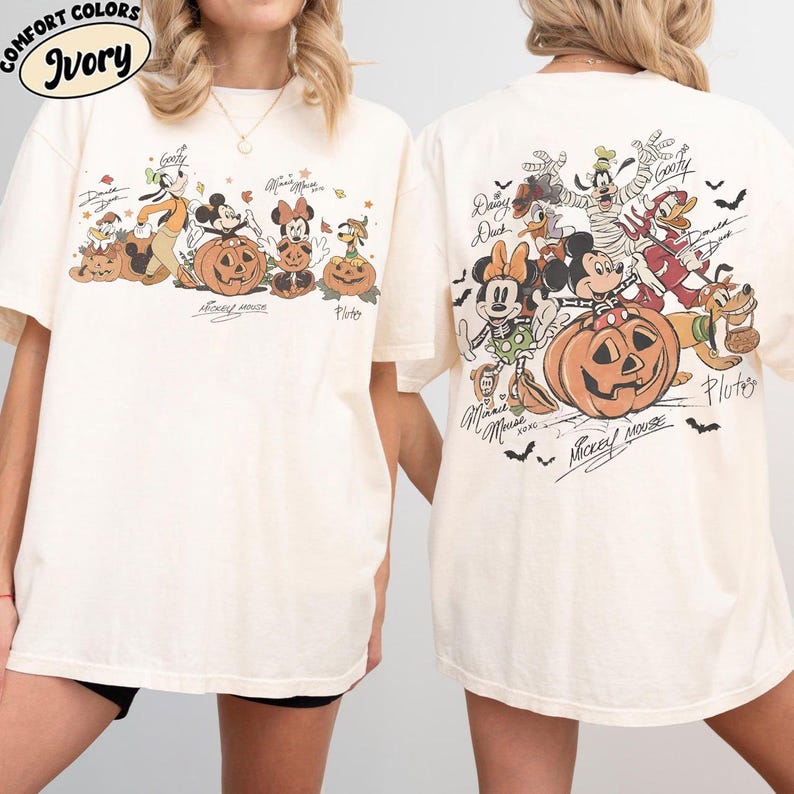May include: Ivory-colored t-shirt featuring a Halloween-themed graphic of Disney characters, including Mickey Mouse, Minnie Mouse, and Goofy, with pumpkins. The front and back of the shirt are shown. The text "Comfort Colors" and "Ivory" are at the top left.