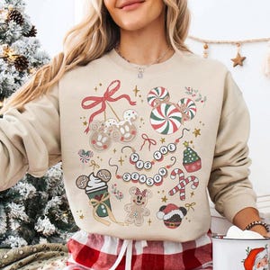 May include: Beige sweatshirt with a festive Christmas design. The design includes gingerbread cookies, candy canes, and the phrase "Tis the Season" in a beaded necklace style. The sweatshirt has a crew neck.