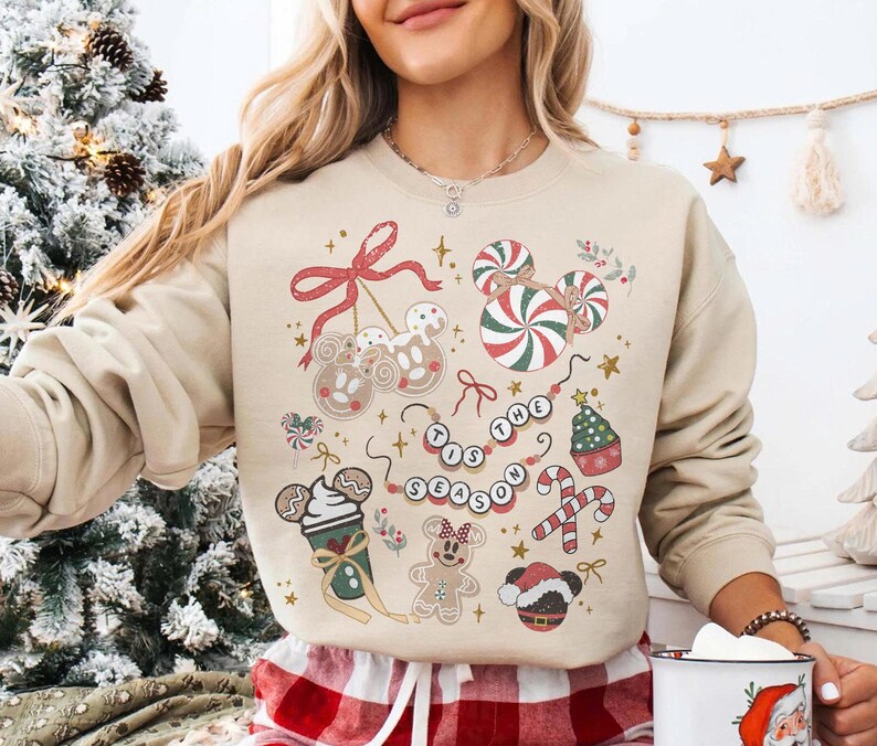 May include: Beige sweatshirt with a festive Christmas design. The design includes gingerbread cookies, candy canes, and the phrase "Tis the Season" in a beaded necklace style. The sweatshirt has a crew neck.