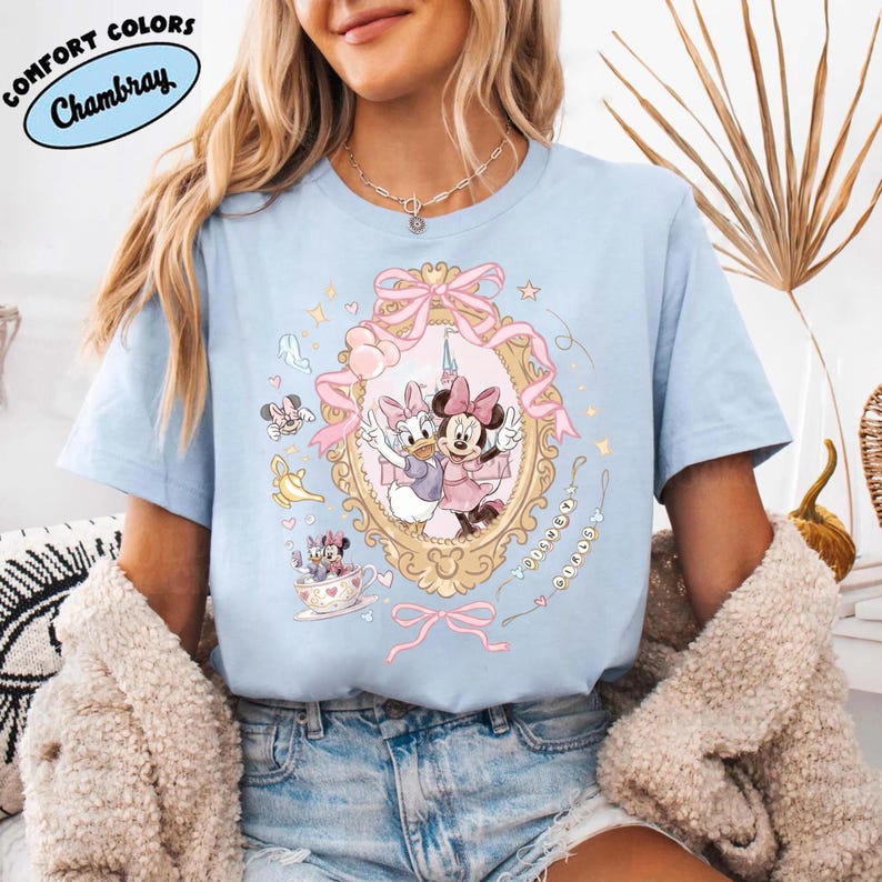 May include: Light blue t-shirt with a cartoon graphic of Minnie Mouse and Daisy Duck in a gold frame with pink bows. The shirt has the words "COMFORT COLORS" and "Chambray" in a blue oval. The design includes hearts, stars, and a teacup.