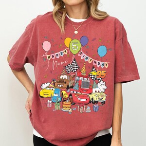 K&ouml;nnte beinhalten: R&ouml;tliches T-Shirt mit einer bunten Grafik von Cartoon-Autos, die einen Geburtstag feiern. Das Design umfasst Luftballons, die Zahl 5 und die W&ouml;rter "Happy Birthday" und "Name". Die Zahl 95 ist ebenfalls sichtbar.