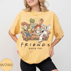 May include: A mustard yellow short-sleeve t-shirt featuring a graphic print of various animated characters sitting on a couch. Below the characters, the text reads "FRIENDS SINCE 1995" in a dark brown font. The shirt has a crew neckline and a loose fit.