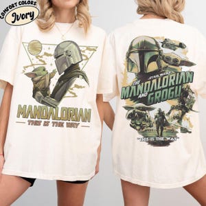 May include: Ivory-colored t-shirt featuring a graphic of The Mandalorian and Grogu. The front design shows the Mandalorian and Grogu with the text "MANDALORIAN - THIS IS THE WAY". The back design includes the text "STAR WARS THE MANDALORIAN AND GROGU".