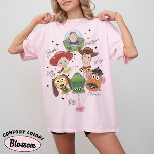 Puede incluir: Una camiseta rosa claro con un gr&aacute;fico de personajes de Toy Story, incluyendo a Woody, Buzz Lightyear y Jessie. La camiseta tiene las palabras "Comfort Colors" y "Blossom" impresas en la parte inferior izquierda.