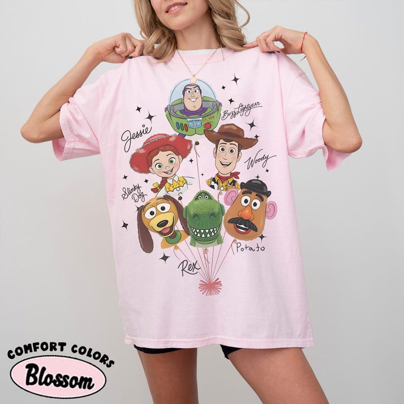 Puede incluir: Una camiseta rosa claro con un gr&aacute;fico de personajes de Toy Story, incluyendo a Woody, Buzz Lightyear y Jessie. La camiseta tiene las palabras "Comfort Colors" y "Blossom" impresas en la parte inferior izquierda.