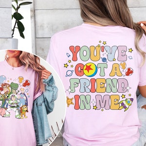 May include: Light pink t-shirt featuring a colorful graphic of Toy Story characters and the phrase "You've Got a Friend in Me." The design includes Woody, Buzz Lightyear, and other characters from the movie, with stars and planets.