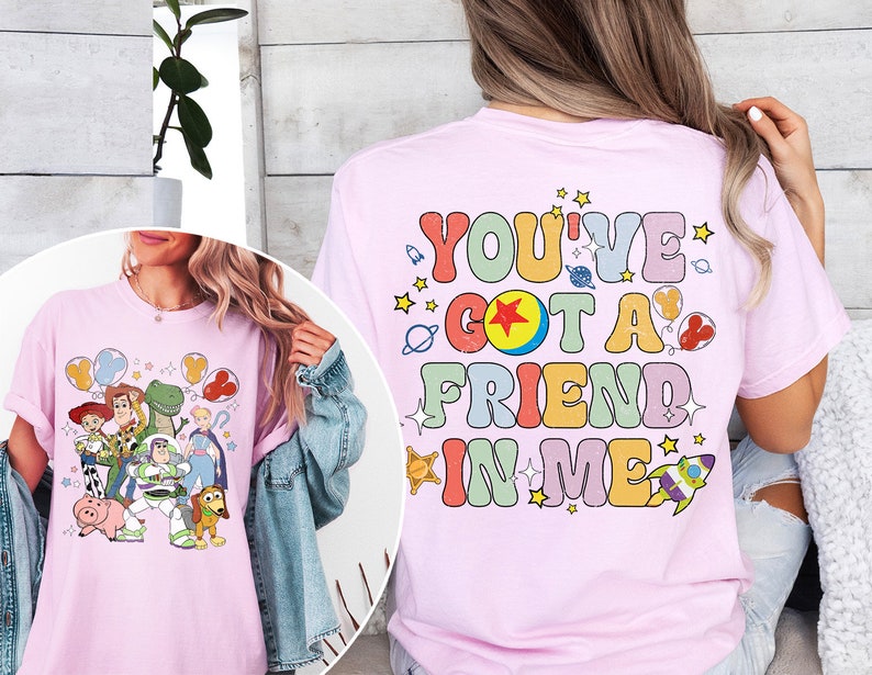 May include: Light pink t-shirt featuring a colorful graphic of Toy Story characters and the phrase "You've Got a Friend in Me." The design includes Woody, Buzz Lightyear, and other characters from the movie, with stars and planets.