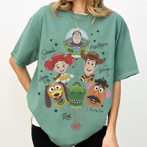 Puede incluir: Camiseta verde salvia con un gr&aacute;fico de personajes de Toy Story como globos. El dise&ntilde;o incluye a Buzz Lightyear, Woody, Jessie, Slinky Dog, Rex y Mr. Potato. Los nombres de los personajes est&aacute;n escritos encima.