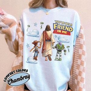 May include: Pale blue Comfort Colors Chambray sweatshirt featuring a graphic of Jesus, Woody, and Buzz Lightyear. The text "You've Got a Friend in Me" is displayed above the characters. The design includes clouds, stars, and parachutes.
