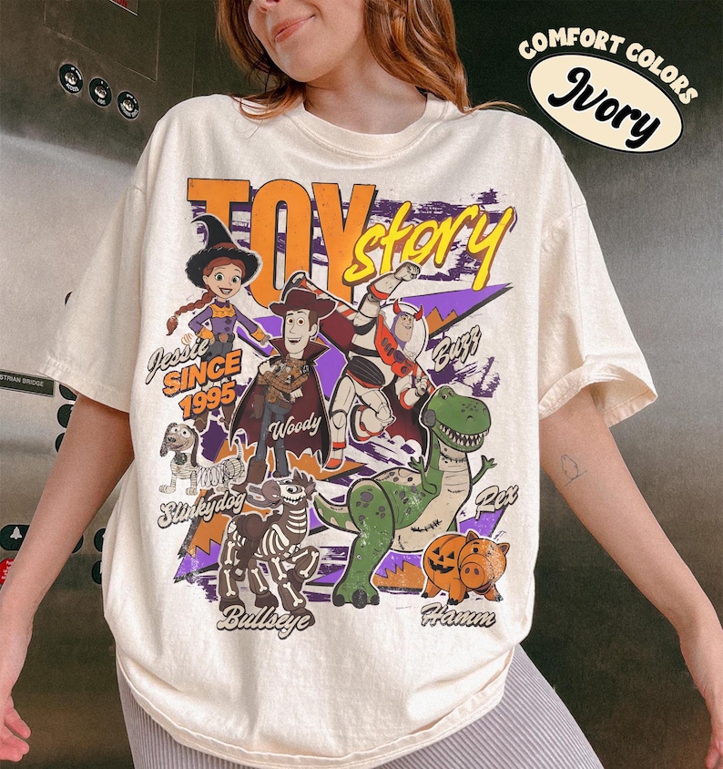 May include: Ivory-colored oversized t-shirt featuring a graphic of Toy Story characters. The design includes Woody, Buzz Lightyear, Jessie, Rex, Hamm, Bullseye, and Slinky Dog. Text includes "Toy Story" and "Since 1995".