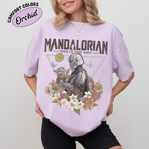 May include: A light purple t-shirt featuring a graphic of The Mandalorian and Grogu surrounded by flowers and butterflies. The text "MANDALORIAN - THIS IS THE WAY" is printed above the image. The shirt also has the text "COMFORT COLORS - Orchid" printed on it.