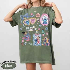 May include: Moss green t-shirt featuring a colorful graphic design with characters from the animated movie Lilo & Stitch. The design includes the text "Lilo & Stitch" and various illustrations of the characters. The shirt is a Comfort Colors brand.