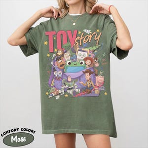 Op de afbeelding: Een mosgroen T-shirt met korte mouwen en een grote grafische print met de tekst "TOY story" en diverse animatiefiguren, waaronder Buzz Lightyear, Woody en een groene kikker, die uit een paarse tas komen met verspreide gele sterren.