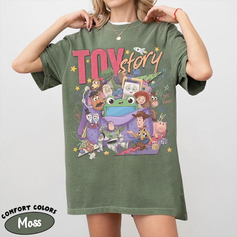 Op de afbeelding: Een mosgroen T-shirt met korte mouwen en een grote grafische print met de tekst "TOY story" en diverse animatiefiguren, waaronder Buzz Lightyear, Woody en een groene kikker, die uit een paarse tas komen met verspreide gele sterren.