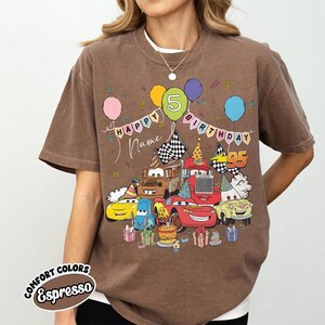 K&ouml;nnte beinhalten: Eine wei&szlig;e Keramik-Tasse mit schwarzem Henkel und dem Text "But First, Coffee" in fetter, schwarzer Schrift. Die Tasse steht vor einem verschwommenen Hintergrund, der eine gem&uuml;tliche Umgebung suggeriert.