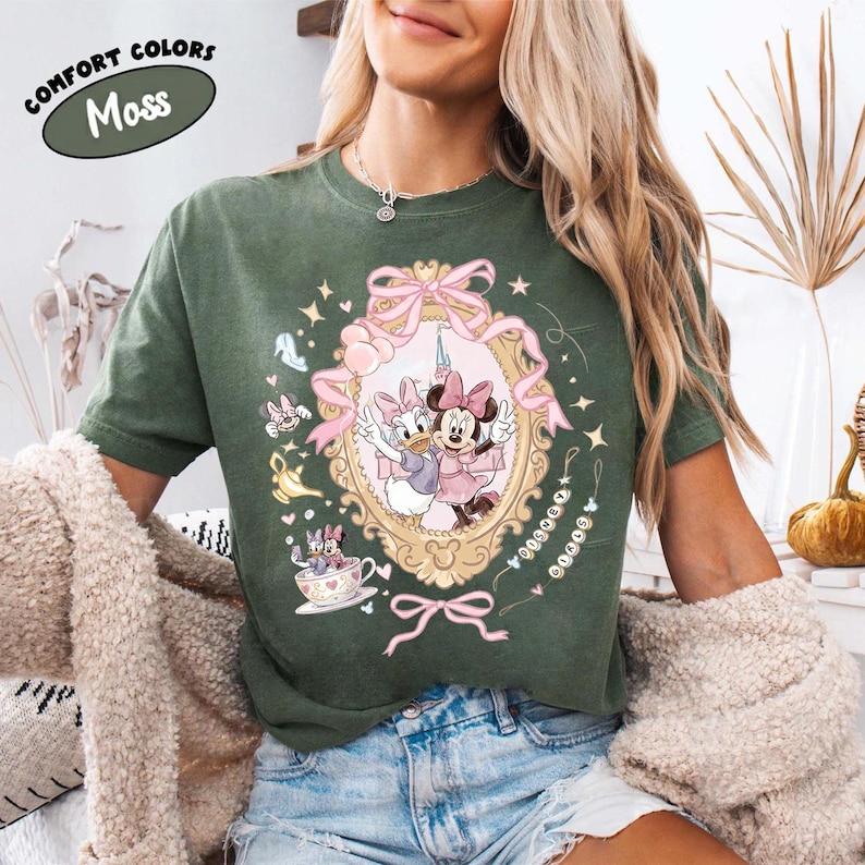 May include: Moss green t-shirt featuring a framed illustration of Minnie Mouse and Daisy Duck in pink dresses. The shirt has the text "COMFORT COLORS" and "Moss" in a circular design. Additional cartoon elements and pink bows decorate the shirt.