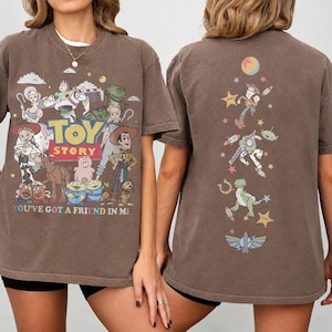 Op de afbeelding: Een bruin t-shirt met korte mouwen en Toy Story-personages op de voor- en achterkant. De voorkant toont het Toy Story-logo met personages zoals Woody, Buzz Lightyear en Jessie, plus de tekst You've Got A Friend In Me. De achterkant toont een verticale rangschikking van personages en sterren.