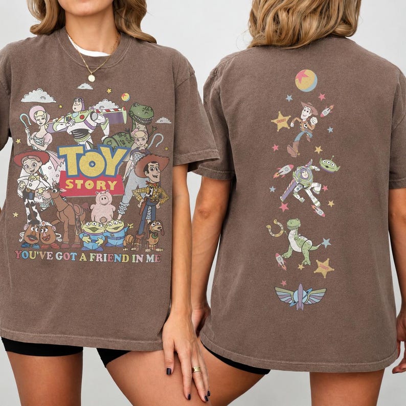 Op de afbeelding: Een bruin t-shirt met korte mouwen en Toy Story-personages op de voor- en achterkant. De voorkant toont het Toy Story-logo met personages zoals Woody, Buzz Lightyear en Jessie, plus de tekst You've Got A Friend In Me. De achterkant toont een verticale rangschikking van personages en sterren.